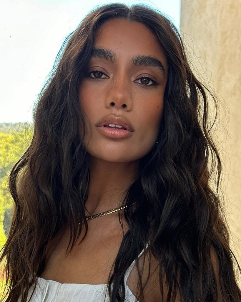 Mulher de pele preta, com cabelo escuro ondulado, acessórios em dourado e blush pêssego 