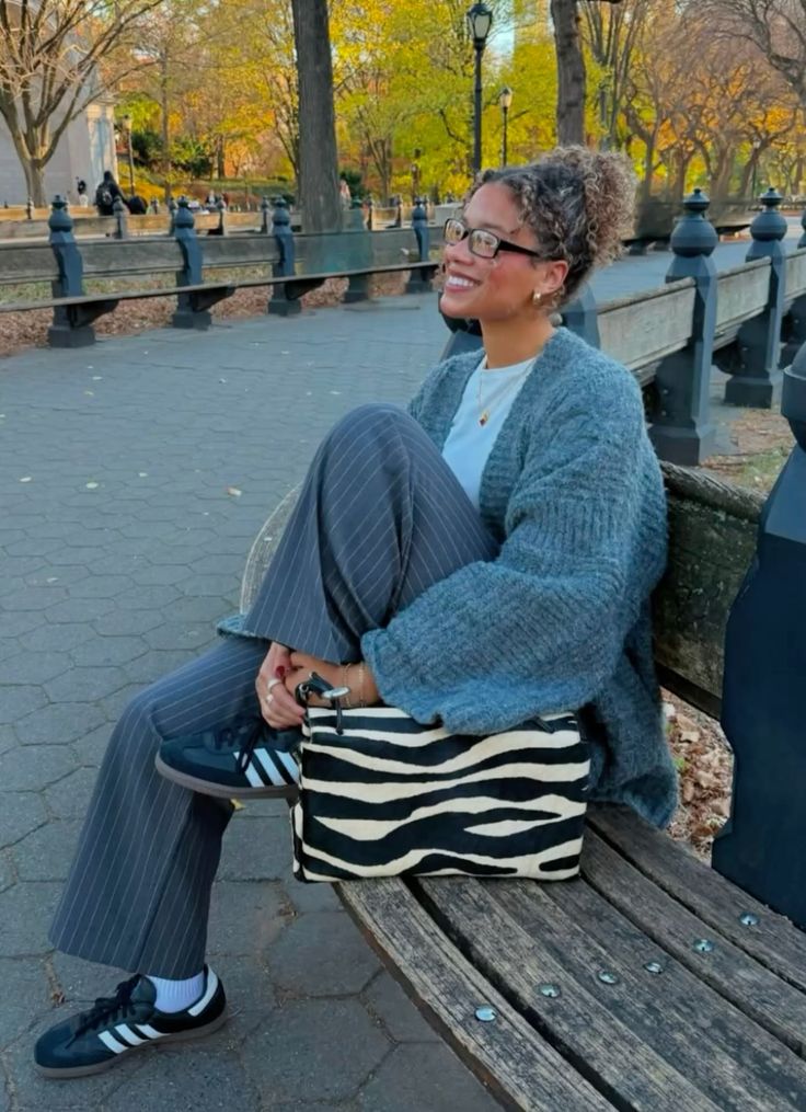Mulher de pele preta e magra, sentada em um banco, com look sóbrio e bolsa de zebra
