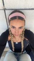 Mulher de pele clara, com roupa escura, acessórios em prata e dourado, tiara ros e cabelo loiro preso em trança