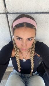 Mulher de pele clara, com roupa escura, acessórios em prata e dourado, tiara ros e cabelo loiro preso em trança