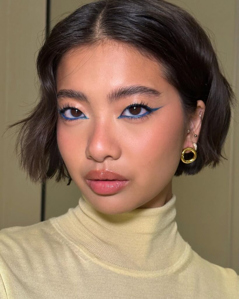Mulher com descendência oriental, blusa de fola alta, cabelo escuro bob, acessórios em dourado e maquiagem com delineado invertido azul 