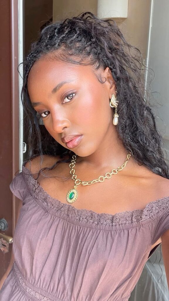 Mulher de pele preta, com blusa arroxeada, cabelo escuro cacheado, acessórios em dourado e maquiagem glow