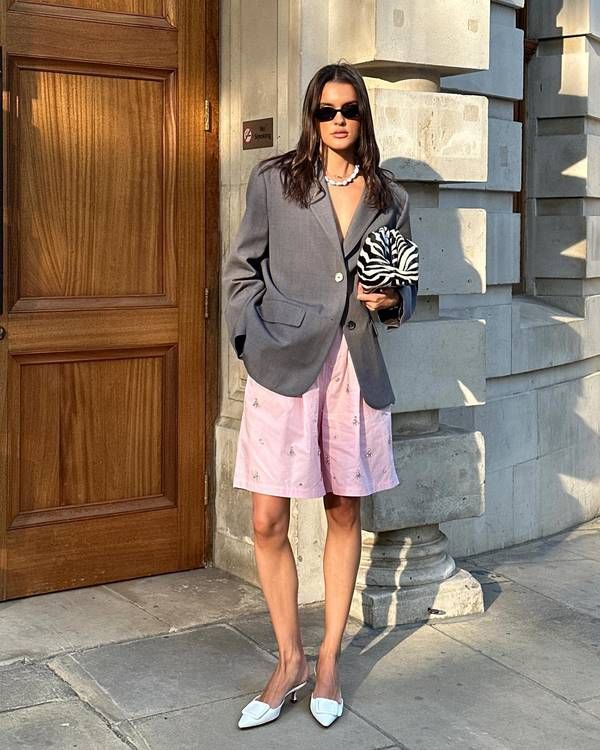 Mulher magra e de pele clara, com blazer cinza, bermuda rosa e bolsa de mão com estampa de zebra