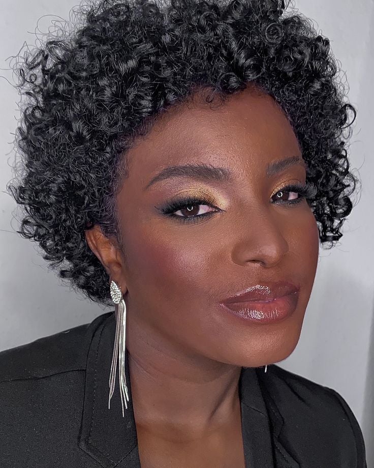 Mulher de pele negra, com cabelo cacheado curto, brinco prata e maquiagem com olhos marcados 