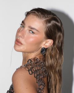Mulher de pele clara, com vestido metalizado, cabelo loiro ondulado e maquiagem glow