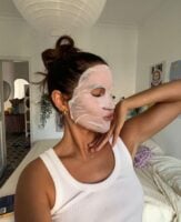 Mulher magra e de pele clara, com cabelo escuro preso, regata branca, acessórios em dourado e máscara de pano no rosto