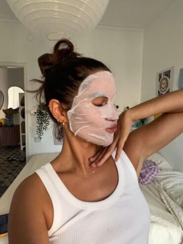 Mulher magra e de pele clara, com cabelo escuro preso, regata branca, acessórios em dourado e máscara de pano no rosto