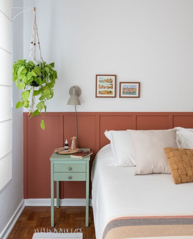 Quarto de casal com cama, cabeceira em madeira ripada, mesa de cabeceira verde, quadros, planta, luminária e pintura em meia parede rosa