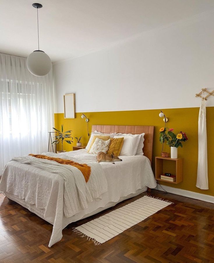 Quarto de casal com cama, cabeceira, quadros, flores, luminária e pintura em meia parede amarela e branca