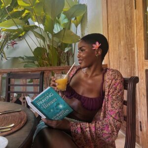 Mulher de pele preta, com biquini vinho, kimono florido. Ela está sentada, tomando suco e lendo