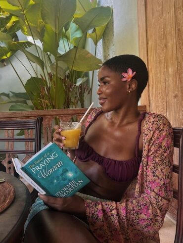 Mulher de pele preta, com biquini vinho, kimono florido. Ela está sentada, tomando suco e lendo