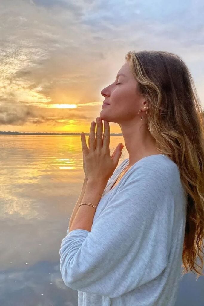 Gisele Bündchen com cabelo loiro solta, blusa cinza, fazendo pose de agradecimento