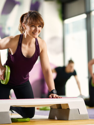Mulher com regata e legging apoiada em um step com uma anilha na mão