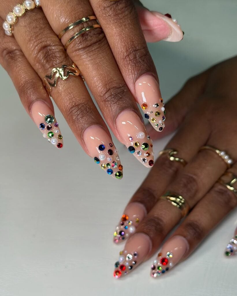 Mãos de pele preta, com anéis em dourado e unha amendoada nude com charms coloridos