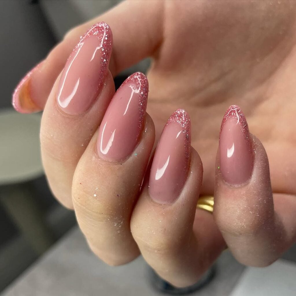 Mão de pele clara, com anel em dourado e unha amendoada rosa-claro com francesinha de glitter
