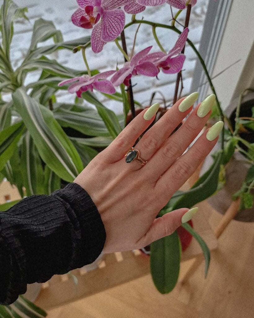 Mão de pele clara, com anel dourado e verde e unha amendoada verde-claro