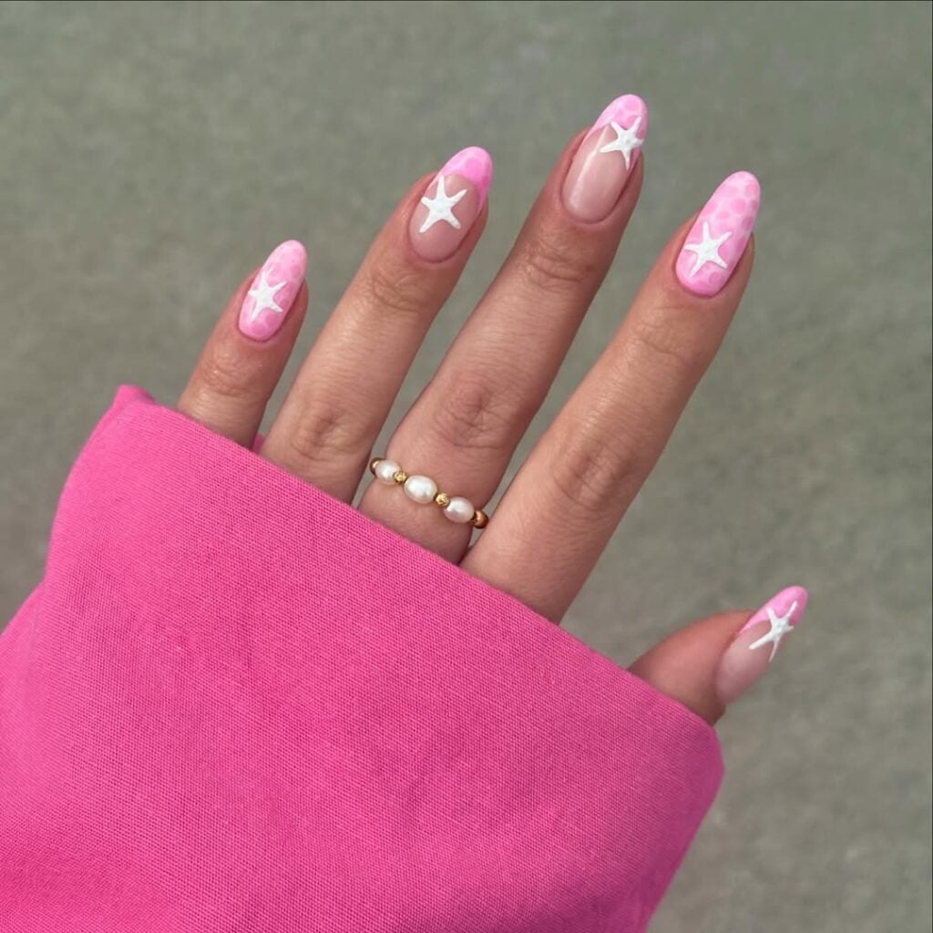 Mão de pele clara, com anel dourado de pérolas, com unha amendoada rosa com estrelas do mar