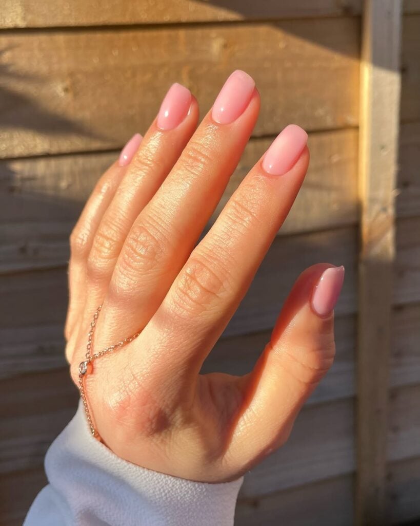 Mão de pele clara, com acessório em dourado e unha curta e quadrada nude com fundo rosado
