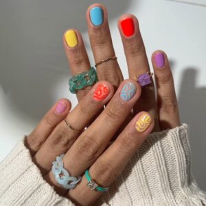 Mão de pele clara, com anéis dourados e coloridos e unhas curtinhas com decorações em neon