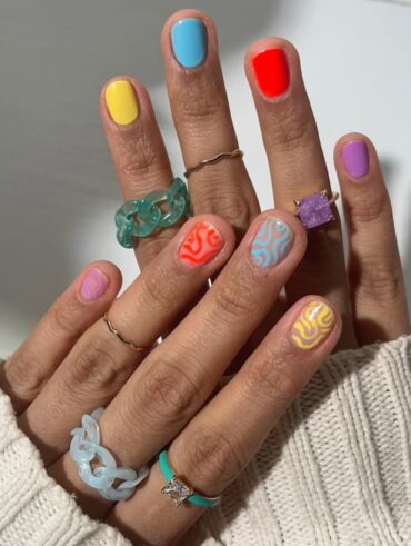 Mão de pele clara, com anéis dourados e coloridos e unhas curtinhas com decorações em neon