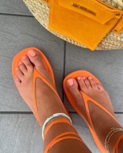 Pés de pele clara, com chinelo de amarração laranja e unha decorada laranja
