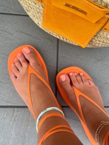 Pés de pele clara, com chinelo de amarração laranja e unha decorada laranja