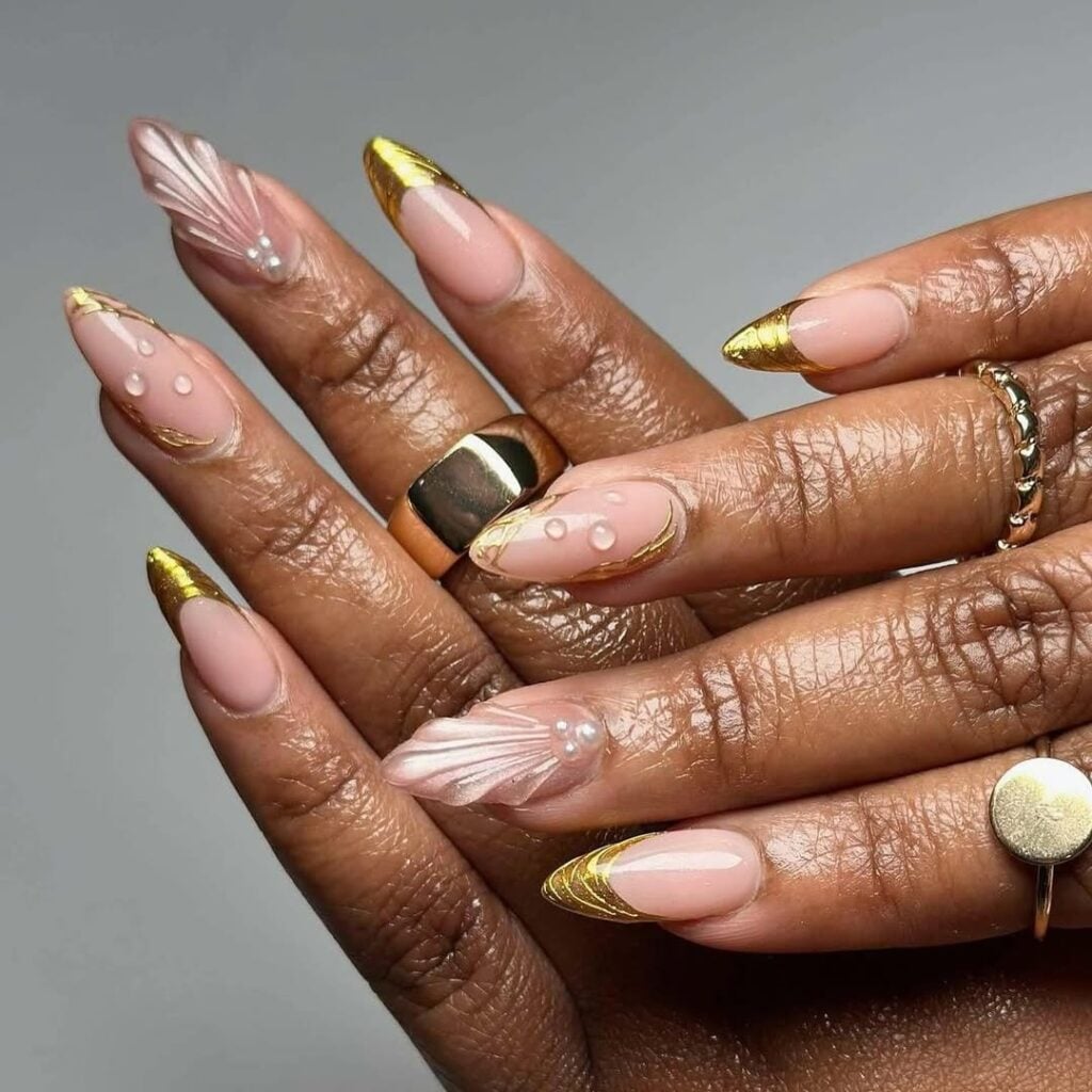 Mãos de pele preta, com anéis em dourado e unha amendoada nude com detalhes em dourado e com gel