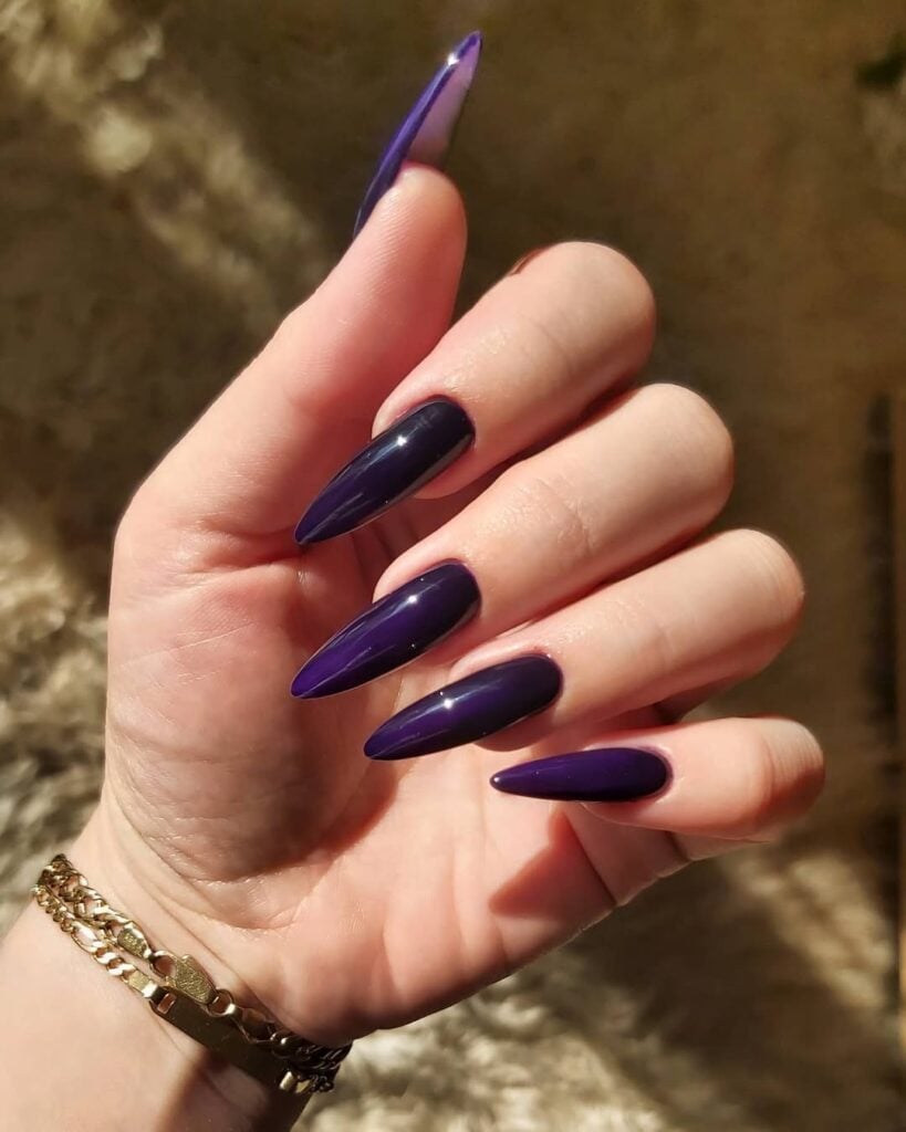 Mão de pele clara, com acessórios em dourado e unha longa com formato stiletto e unha roxo-escuro