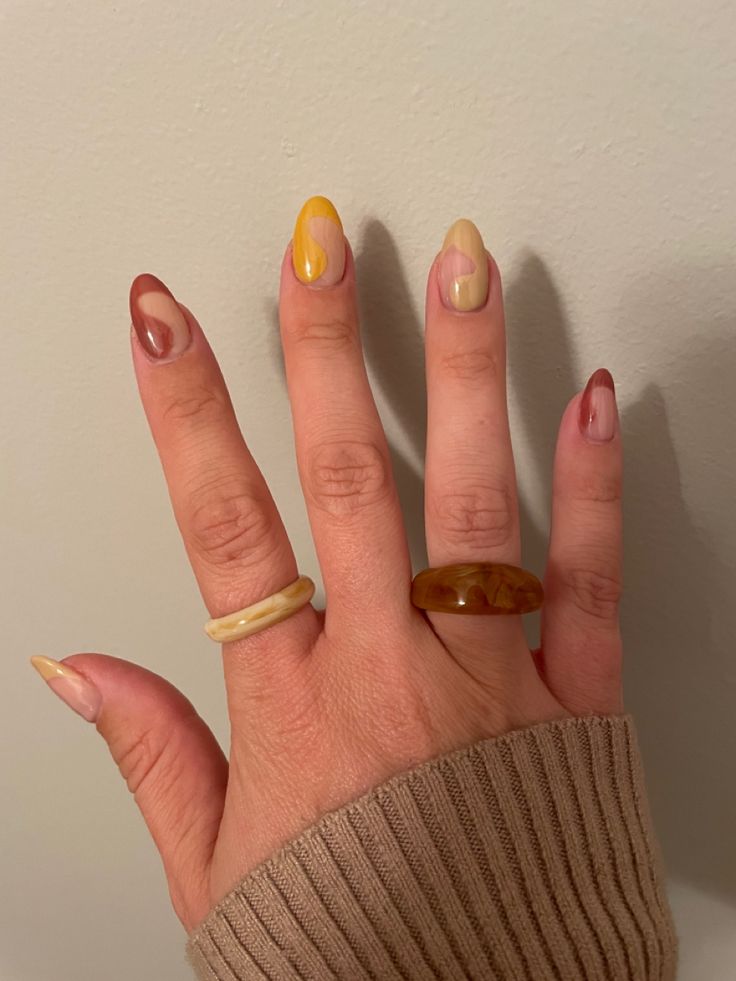 Mão de pele clara, com anéis coloridos e unhas amendoadas com ondulações terracota
