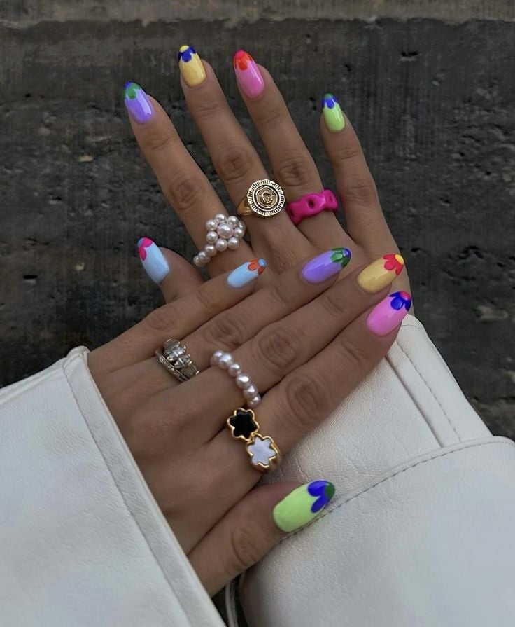 Mãos de pele clara, com anéis diversos e unhas amendoadas coloridas com flores nas pontas