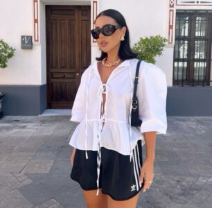 Mulher jovem e magra com pele clara e cabelos escuros. Ela usa blusa branca com manga bufante, bermuda atlética, óculos de sol, colar e brincos.