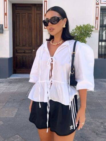 Mulher jovem e magra com pele clara e cabelos escuros. Ela usa blusa branca com manga bufante, bermuda atlética, óculos de sol, colar e brincos.