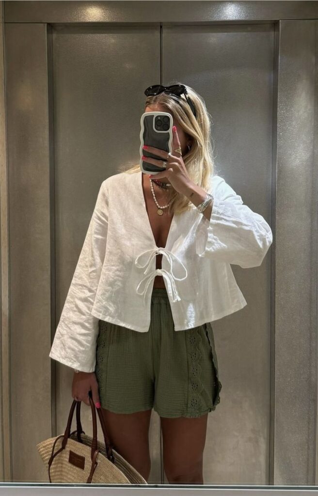 Mulher jovem e magra na frente do espelho. Ela veste blusa branca com amarração e bermuda verde.