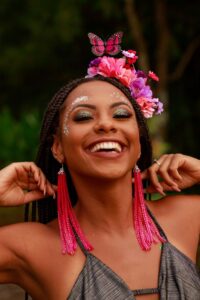 Mulher jovem de pele negra com tiara de flores rosas e roxas e brinco rosa comprido