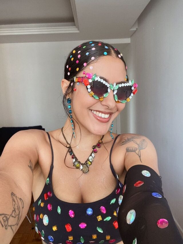 Mulher de pele clara, com top preto com pedras, óculos decorado e cabelo escuro decorado com adereços