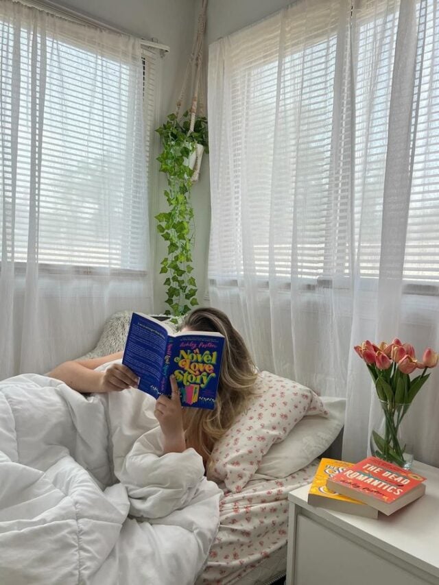 Mulher de pele clara, com cabelo loiro, deitada com um livro aberto na frente do rosto