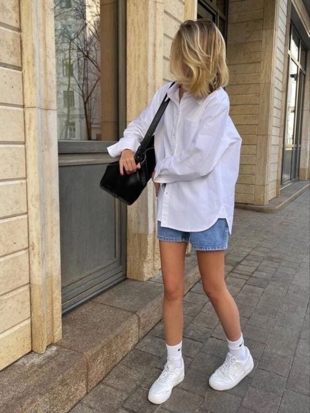 Mulher de pele clara, com short jeans, camisa branca, bolsa preta e tênis branco
