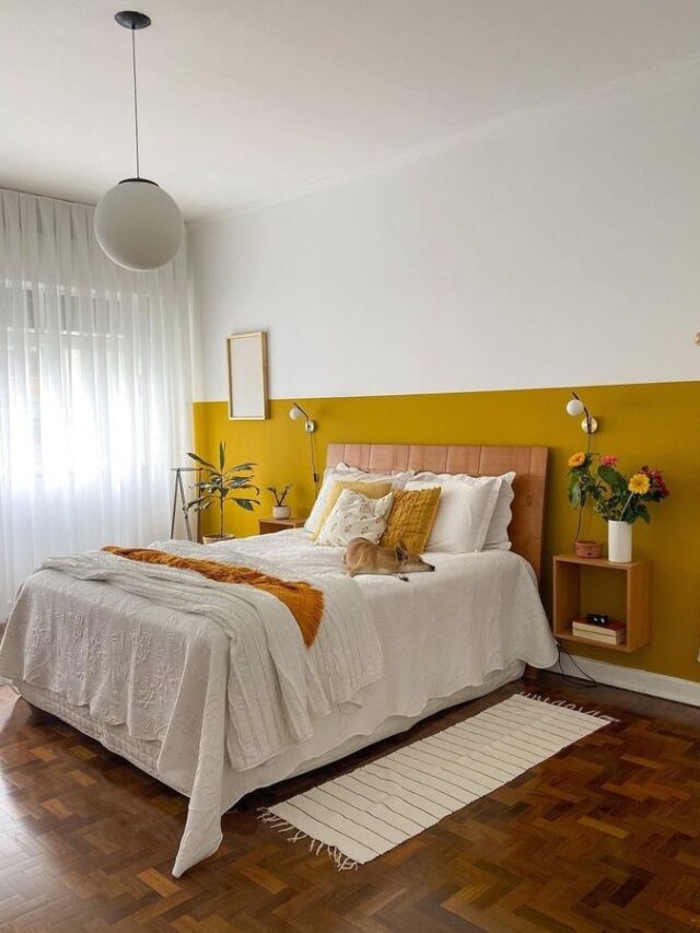 Quarto de casal com cama, cabeceira, quadros, flores, luminária e pintura em meia parede amarela e branca