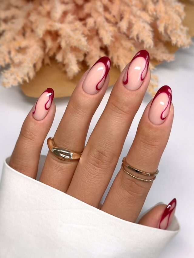 Mão de pele clara, com anéis em dourado e unha amendoada nude com francesinha vermelho-escuro e linhas fluidas