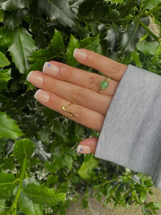 Mão de pele clara, com anéis em dourado, unha curta e quadrada em gel e francesinha branca com detalhe de olho grego