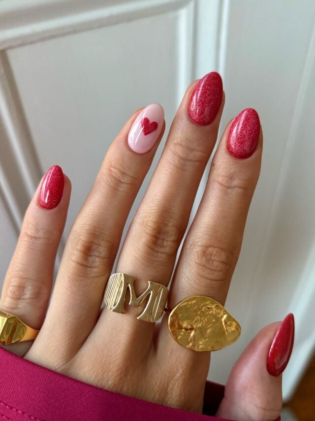 Mão de pele clara, com anéis em amarelo e unha amendoada vermelha com glitter e coração