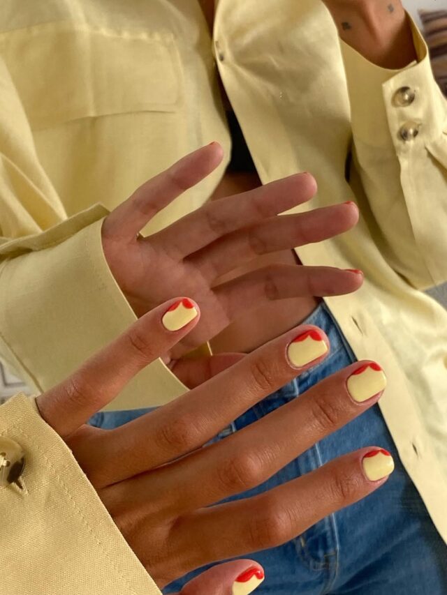 Mão de pele branca, com unha curta e quadrada, com esmalte amarelo e vermelho e técnica de nail art