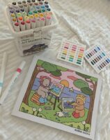 Lençol branco com livro de colorir Bobbie Goods e canetinhas coloridas.