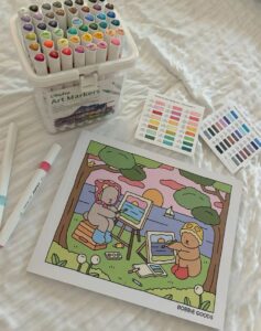Lençol branco com livro de colorir Bobbie Goods e canetinhas coloridas.