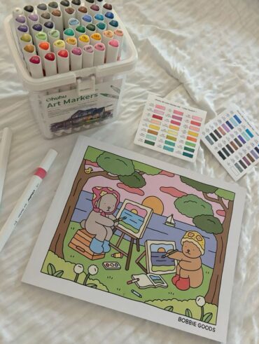 Lençol branco com livro de colorir Bobbie Goods e canetinhas coloridas.
