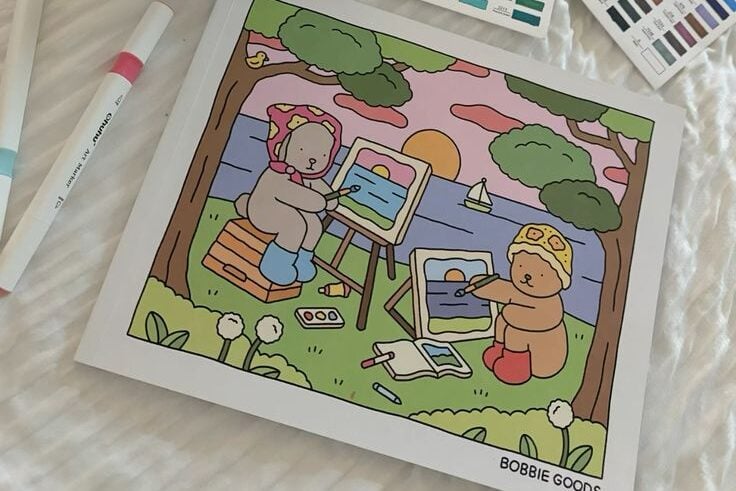 Lençol branco com livro de colorir Bobbie Goods e canetinhas coloridas.