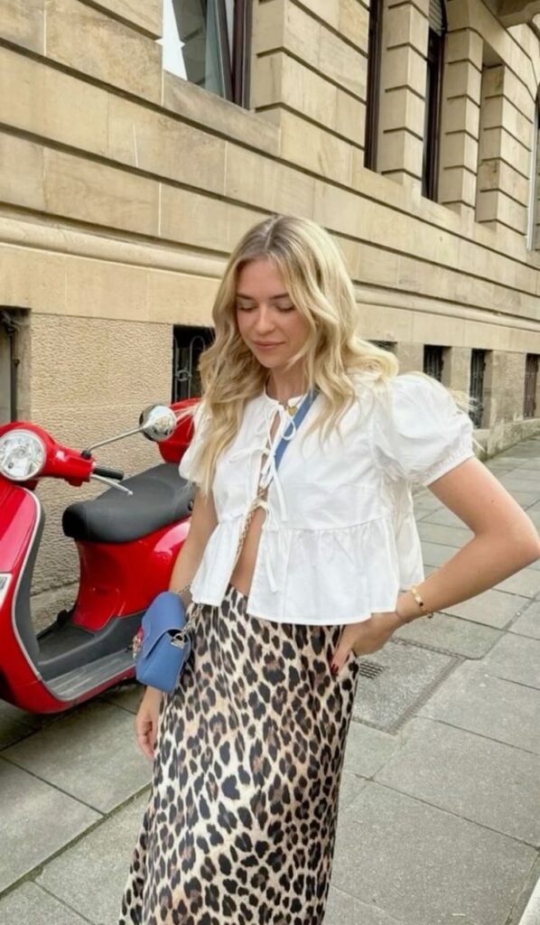 Mulher jovem, magra e loira. Ela veste blusa branca com amarração frontal e mangas bufantes e saia longa com estampa de leopardo.
