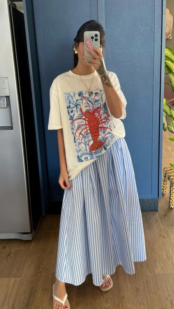 Mulher jovem, magra e de pele clara. Ela está usando uma blusa com estampa, saia longa listrada e chinelo.