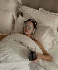 Mulher jovem deitada em uma cama com um copo de vinho na mão, fones de ouvido e skin care no rosto.