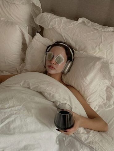 Mulher jovem deitada em uma cama com um copo de vinho na mão, fones de ouvido e skin care no rosto.
